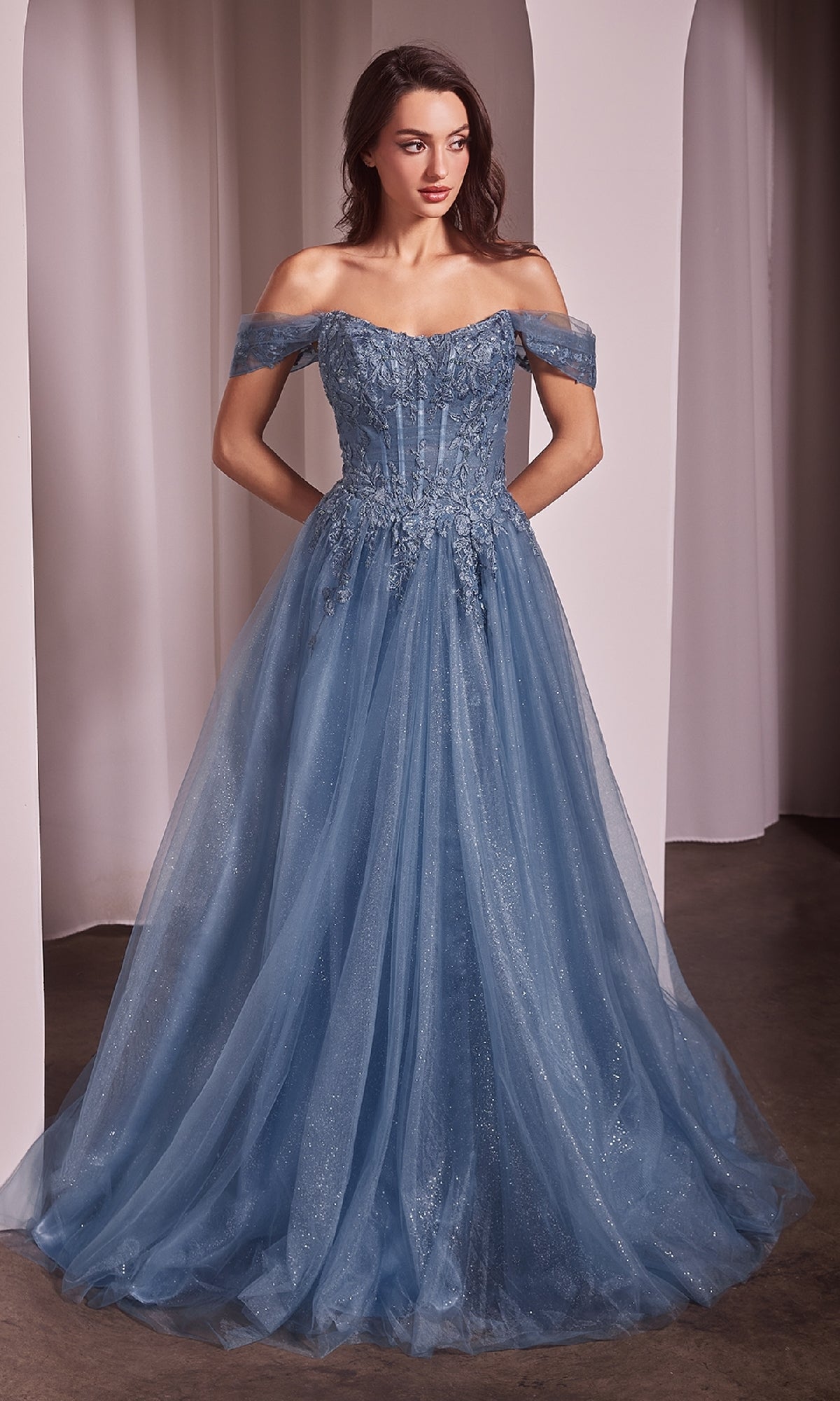 A woman stands indoors wearing the Off-Shoulder Long Prom Dress: Ladivine CD799—a floor-length blue gown with a fitted bodice and tulle skirt. White drapery hangs in the background.
